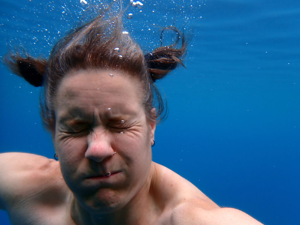 A woman with spacebuns under water. Her eyes and mouth are closed tightly and a few bubbles are escaping